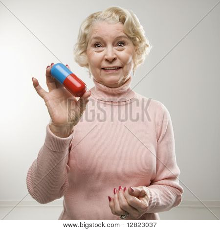 Caucasian senior woman holding oversized pill at viewer.