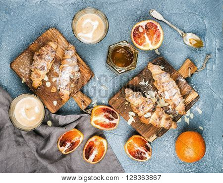 Traditional Italian style home breakfast. Latte in glasses, almond croissants on rustic wooden boards and red bloody Sicilian oranges over concrete textured table, top view, horizontal