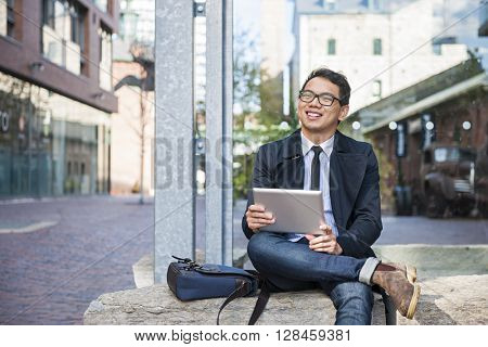 Young smiling asian business man holding a digital tablet sitting outside on city street looking away