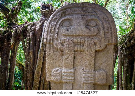 A mysterious statue of a male person  stands in the rainforest next to an old tree with large roots. The statues of San Agustin are a mystery to historians - San Agustin, Colombia in December 2015