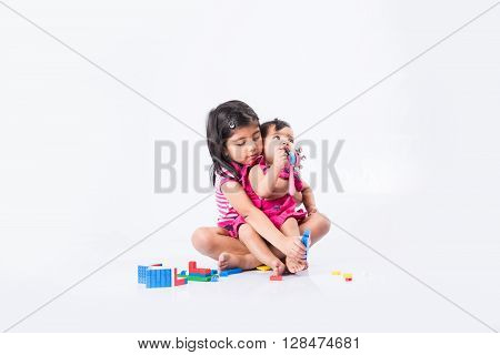 indian small girls or asian small siblings playing with colourful blocks over white background, cute little indian girl baby sitting or taking care of younger sister, two small indian sisters playing