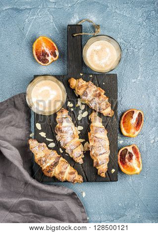 Italian style home breakfast. Latte coffee, almond croissants and red bloody oranges on dark wooden serving  board over concrete textured table, top view