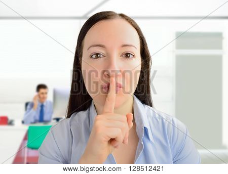 Portrait of a young businesswoman with her finger on her lips at the office with the colleague working in background