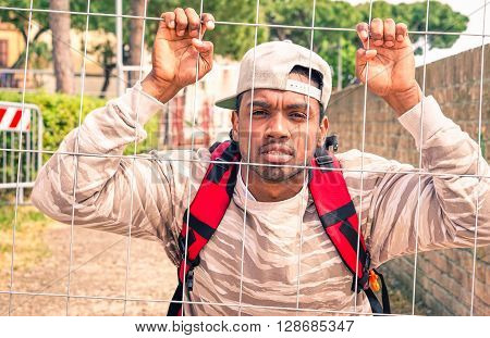 African man refugee clinging to metal net looking camera in pensive mood - Young afro migrant male holding border mesh - Black guy behind bars - Unhappiness cultural and political barriers concept
