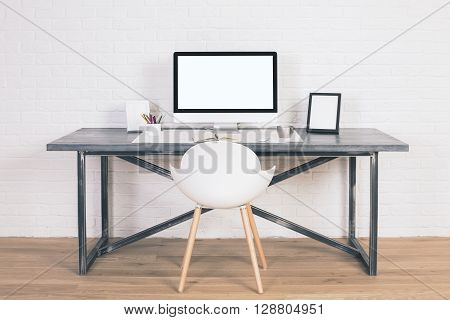 Front view of designer desk with blank white computer screen frames and other items with white chair next to it. Wooden floor and white brick wall background. Mock up