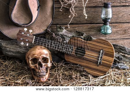Still life human skull and ukulele on hay with traditional leather boots and american west rodeo brown felt cowboy hat background vintage and dark tone for horror halloween