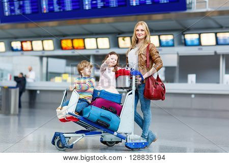 Two adorable little siblings, kid boy and girl sitting on suitcases on international airport. Young mother pushing. Brother and sister, happy family wating for flight and going on vacations.