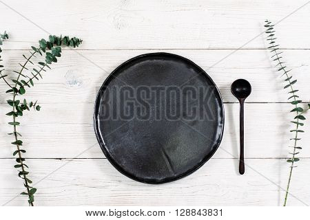 Top view empty black plate and cutlery on rustic wooden table. Ceramic black plate and spoon on white wooden background with free space. Flat lay of handmade black dish on white wooden table.