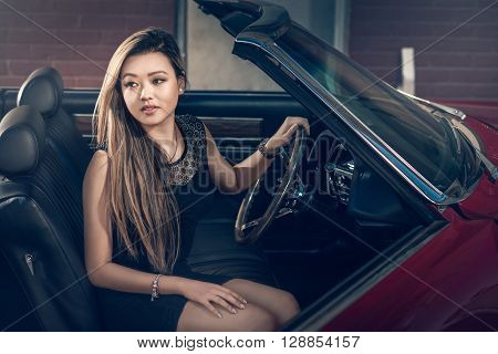 Attractive girl sitting in classic american muscle car parked on the street near the old barn