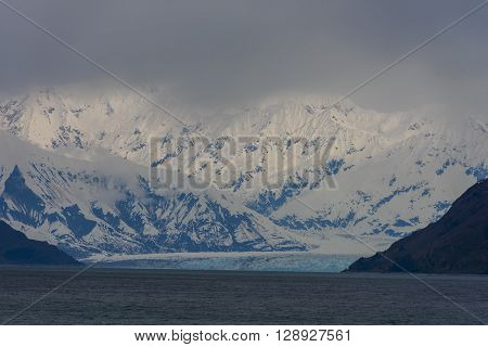 A dramatic photo of a glacier with snow-capped moiuntains in the background
