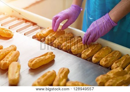Hands in gloves touching eclairs. Eclair shells on narrow conveyor. Employee of confectionery plant. Worker responsible for quality.