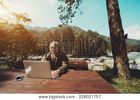 Aged Grey Haired Hunter Is Sitting At Big Wooden Table And Checking His E-mail Via Laptop; Mature Ma