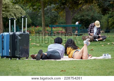 Asian Loving Couple Of Tourists With Suitcases Lies On The Green Grass In The Park. The Champ De Mar