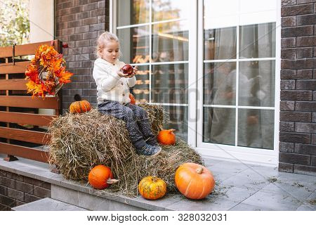 Adorable Smiling Caucasian Baby Toddler In White Knittes Jacket Sitting On The Haystack With Pumpkin