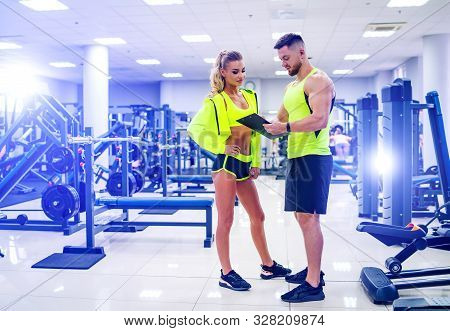 Personal Trainer With A Folder Working With Client Woman At A Gym. Creating A Scedule. Wears Bright 