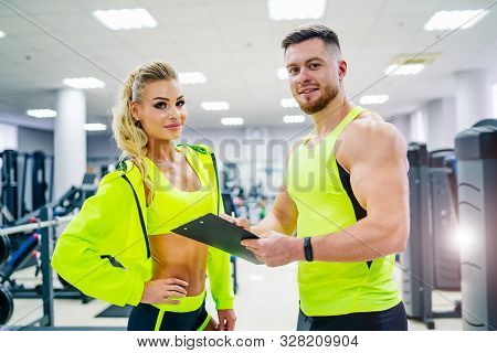 Personal Trainer With A Folder Working With Client Woman At A Gym. Creating A Scedule. Wears Bright 