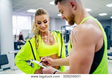 Personal Trainer With A Folder Working With Client Woman At A Gym. Creating A Scedule. Wears Bright 