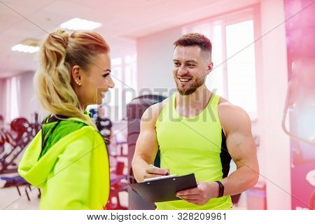 Personal Trainer With A Folder Working With Client Woman At A Gym. Creating A Scedule. Wears Bright 