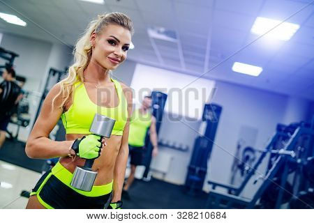 Gorgeous Blonde Woman Is Warming Up With A Dumbbell And Doing Workout At A Gym Looking In Camera. Sh