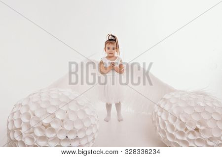 A Girl In A White Dress With Angel Wings Holding On A White Background. An Angel-child Have Big Whit