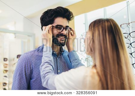 Femaale Ophthalmologist Helping Man To Choose Glasses In Optical Store