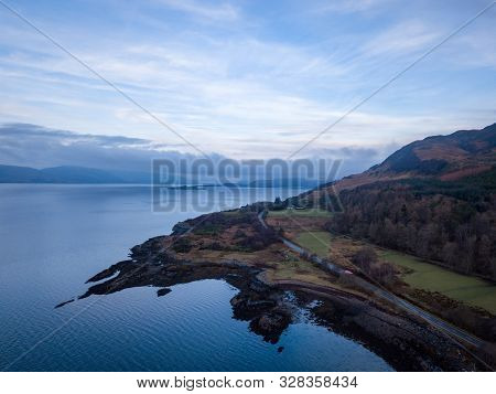 A Road With A Truck And Some Cars Passes Around The Rocky Scottish Coastline With A View Of The Loch