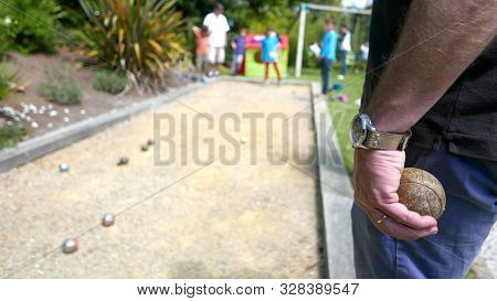 Hand Of A Man Holding A Rust Ball Of Petanque With Family In The Background