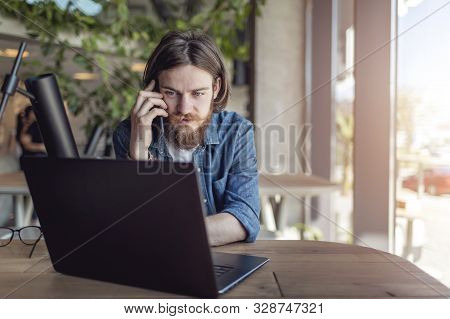 Young Casual Startupper Talking On Mobile Phone While Working By Laptop In A Loft Workspace