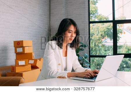 Concentrated Asian Female Startupper In Stylish White Jacket Working With Laptop With Parcel Boxes O