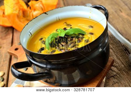 Enamel Pot Of Cream Pumpkin, Carrot Soup With Fresh Basil, Pumpkin Seed And Laddle On Rustic Wooden 