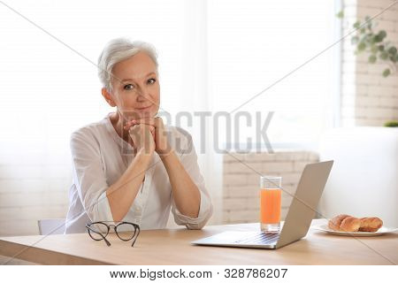 Mature Woman With Laptop Sitting At Table In Kitchen. Smart Aging