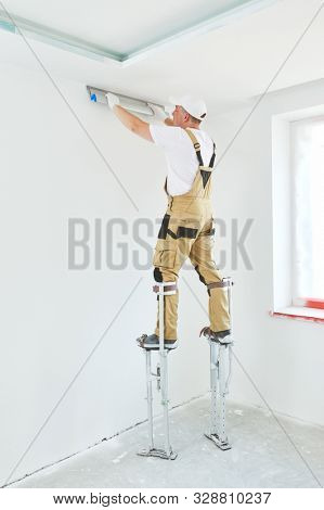 Painter in stilts with putty knife. Plasterer smoothing ceiling surface at home renewal