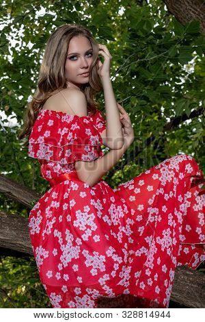 A Beautiful Young Girl With Perfect Skin, With Long Hair In A Red Dress Lies On A Tree Branch With A