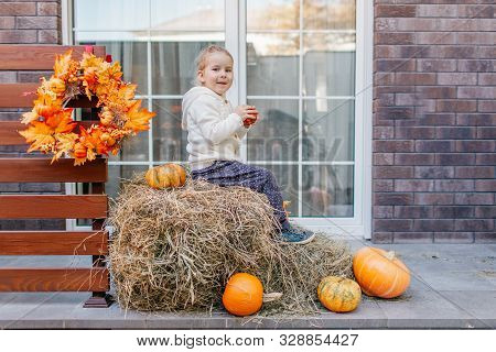 Adorable Blonde Baby Toddler In White Knittes Jacket Sitting On The Haystack With Pumpkins At Porch,