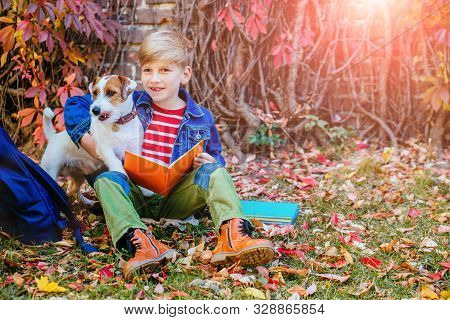 Preteen Cute Boy Sitting On Grass In A Fall Autumn Time Reading His Book, Hugging His Dog Over Climb