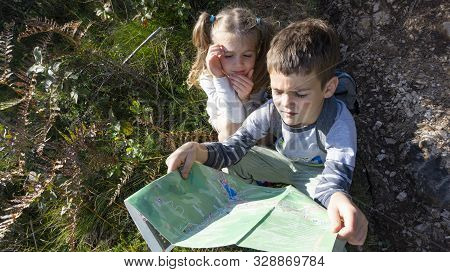 Two Kids With Backpacks Are Looking For A Hiking Trail 
On The Tourist Map In The Mountains In A Tem