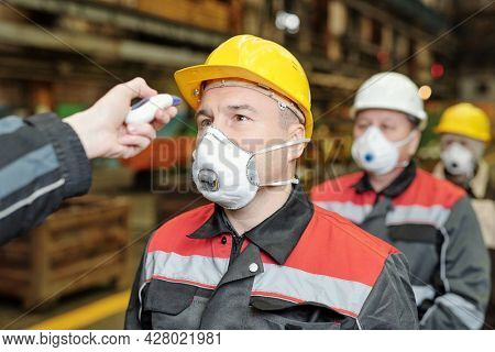 Hand with infrared-thermal thermometry checking temperature of factory workers in masks and helmets standing in line