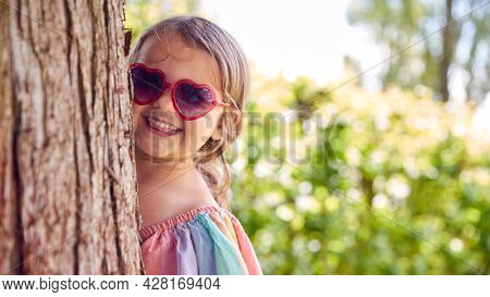 Smiling Young Girl Wearing Sunglasses Playing Hide And Seek Behind Tree In Garden