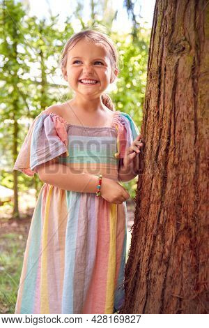Smiling Young Girl Playing Hide And Seek Behind Tree In Garden