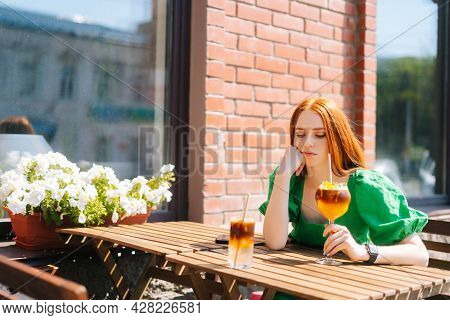 Sad Thoughtful Young Woman Drinking Cocktail Through Straw Sitting At Table In Outdoor Cafe In Sunny