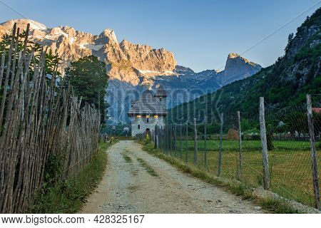 A Path With Wooden Fence Leads The Catholic Church In The Theth Valley, On Background The Mountains 