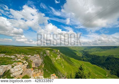 Mountain Gorge On A Clear Sunny Day. Beautiful Clouds, Green Slopes. A Rocky Cliff In The Foreground