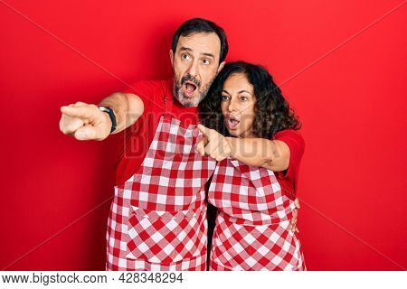 Middle age couple of hispanic woman and man wearing cook apron pointing with finger surprised ahead, open mouth amazed expression, something on the front 