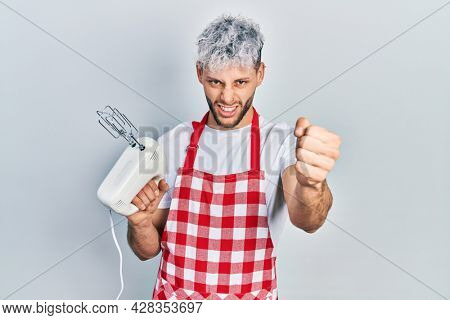 Young hispanic man with modern dyed hair holding food processor mixer machine annoyed and frustrated shouting with anger, yelling crazy with anger and hand raised 