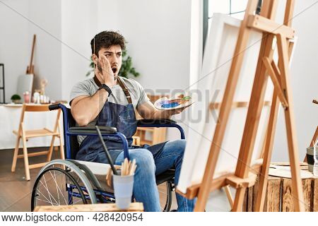 Young hispanic man sitting on wheelchair painting at art studio afraid and shocked, surprise and amazed expression with hands on face 