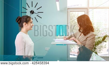 Lady Office Manager In White Blouse Shows Registration Book To Woman Client Standing At Reception In