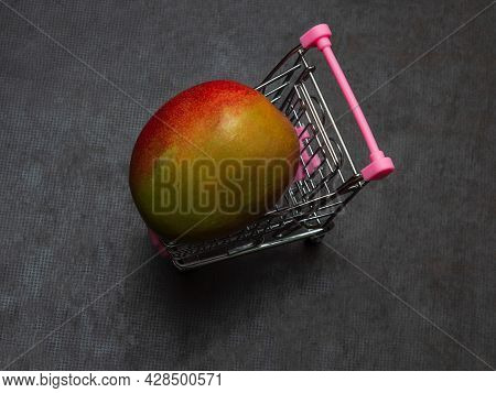 One Green Red Mango Is Lying In A Small Shopping Trolley. An Exotic Tropical Fruit In A Toy Metal Ca