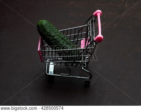 One Green Cucumber Is Lying In A Small Shopping Trolley. A Farmer's Vegetable In A Toy Metal Cart.