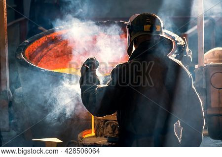 Worker Of Metal And Alloys Foundry At A Metallurgical Plant, Rear View. Smoke From Red Metal