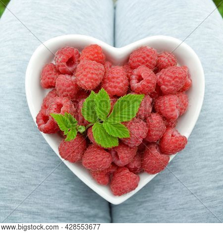 A Woman Farmer Holds A Heart-shaped Plate On Her Lap, In Which There Is A Ripe Raspberry With A Leaf